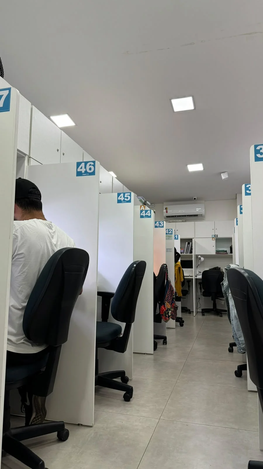 Estudantes em cabines individuais numeradas, com divisórias altas e cadeiras ergonômicas